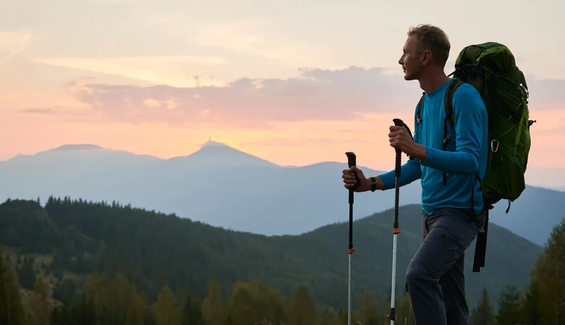 Vandrer med udstyr på toppen af en bakke over for blånende bjergkamme i solnedgang